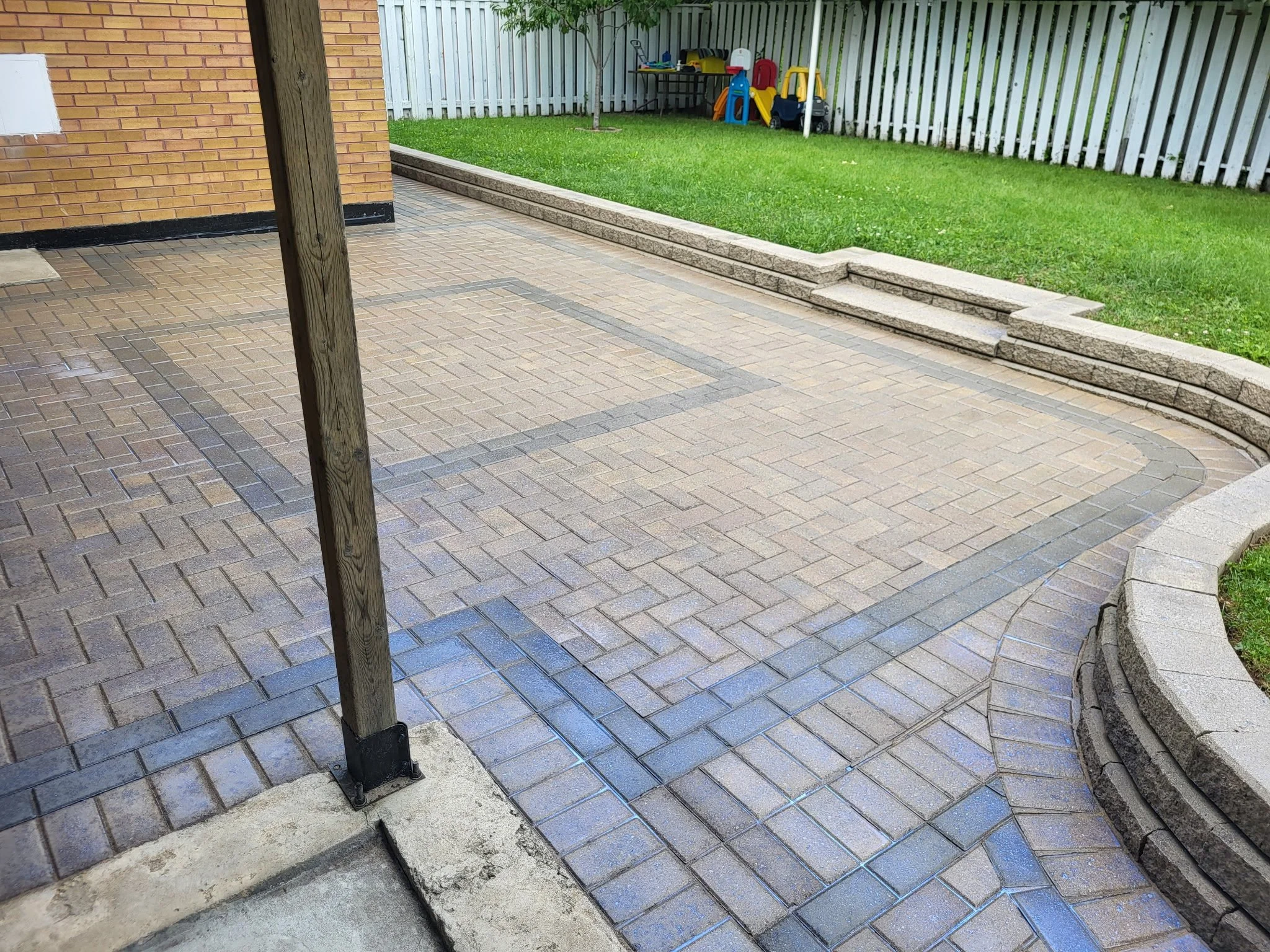 A backyard patio with tan brick paving, a wooden post, concrete steps, a green lawn with a white picket fence, and children's play equipment in the background.