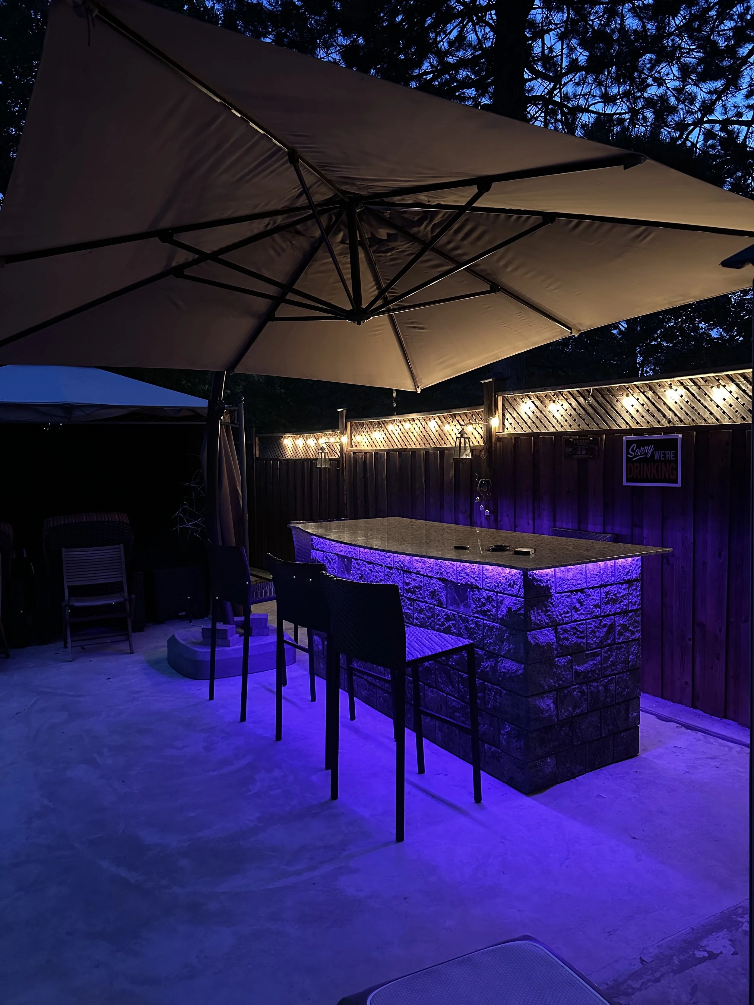 Outdoor patio area at night illuminated by purple and warm string lights, featuring a bar with purple lighting, chairs, large umbrella, and a wooden privacy fence with a sign that says 'Sorry, we're drinking.'