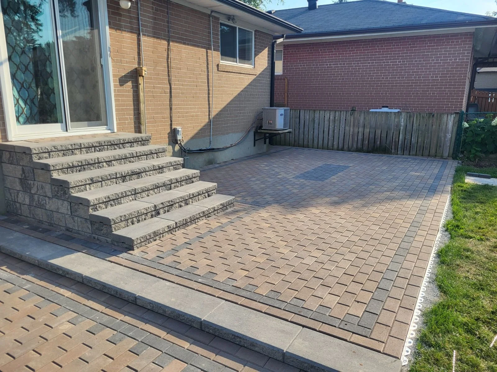 A backyard brick patio with steps leading up to a sliding glass door, surrounded by a lawn and a wooden fence.