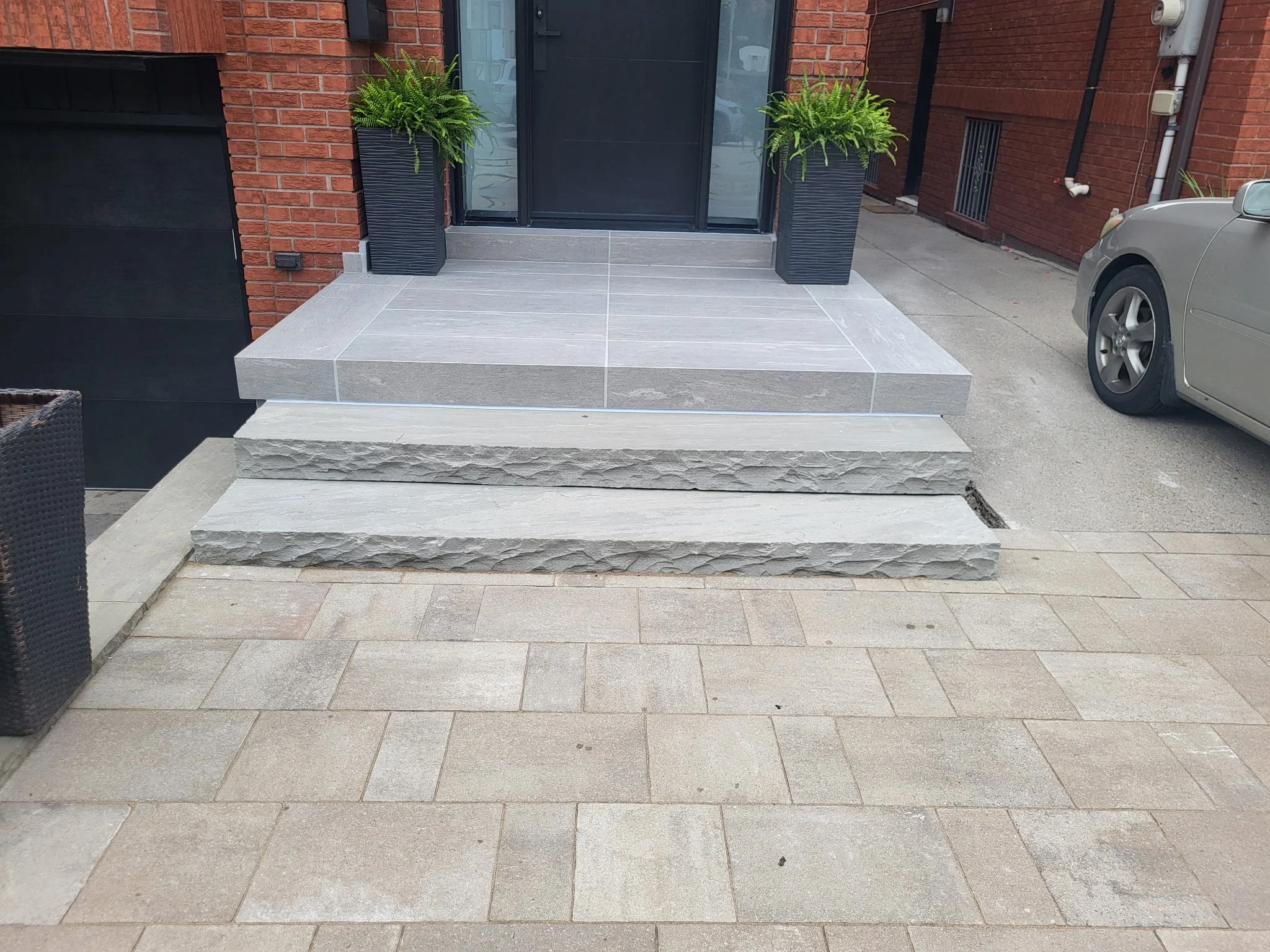 Front porch with large gray tiles, two tall black planters with green ferns, black door with glass, brick walls, parked car on right side.