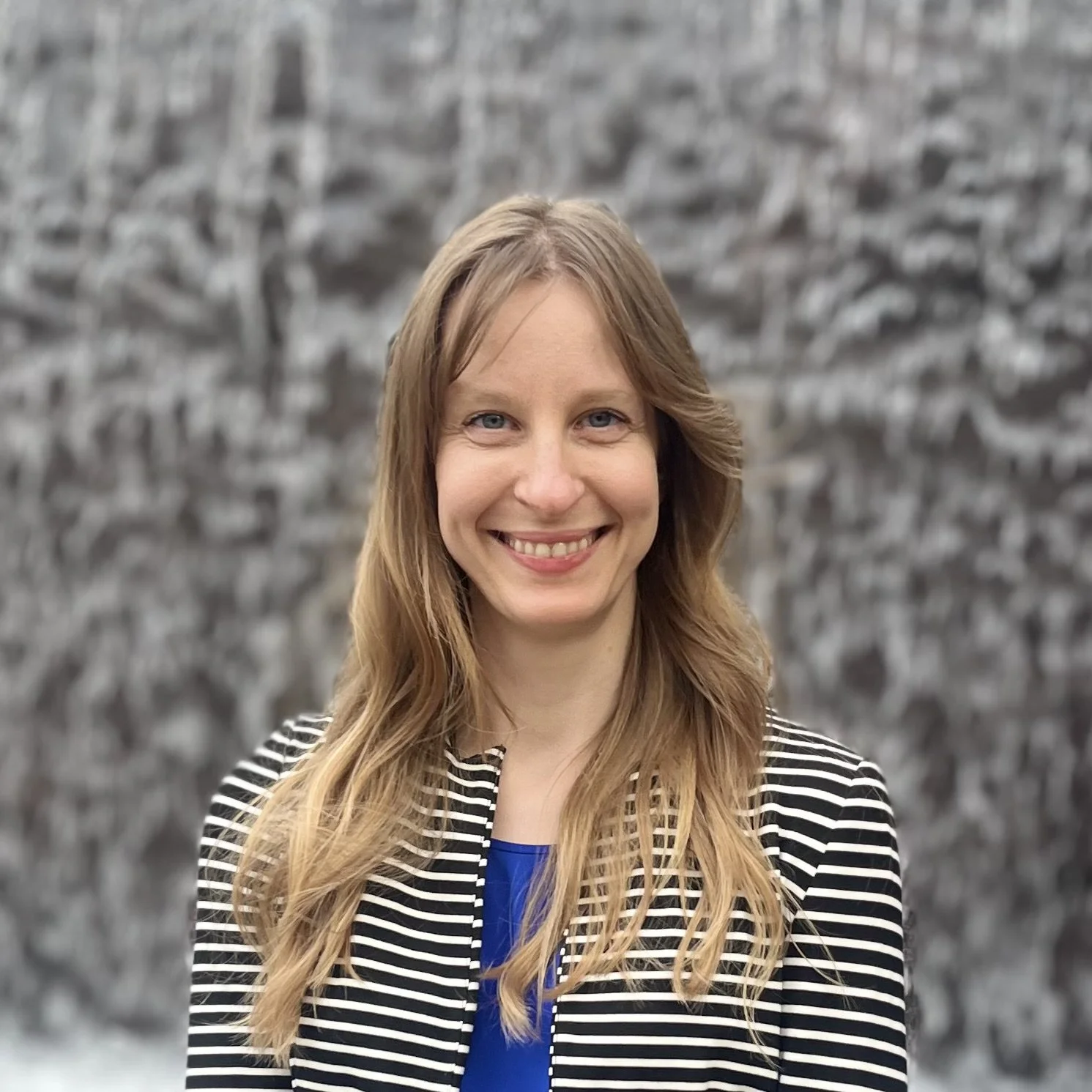 A woman with long, wavy blonde hair smiling in front of a blurred outdoor background with rocks.