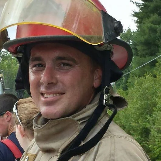 Firefighter wearing a yellow and red helmet and tan uniform, smiling outdoors.