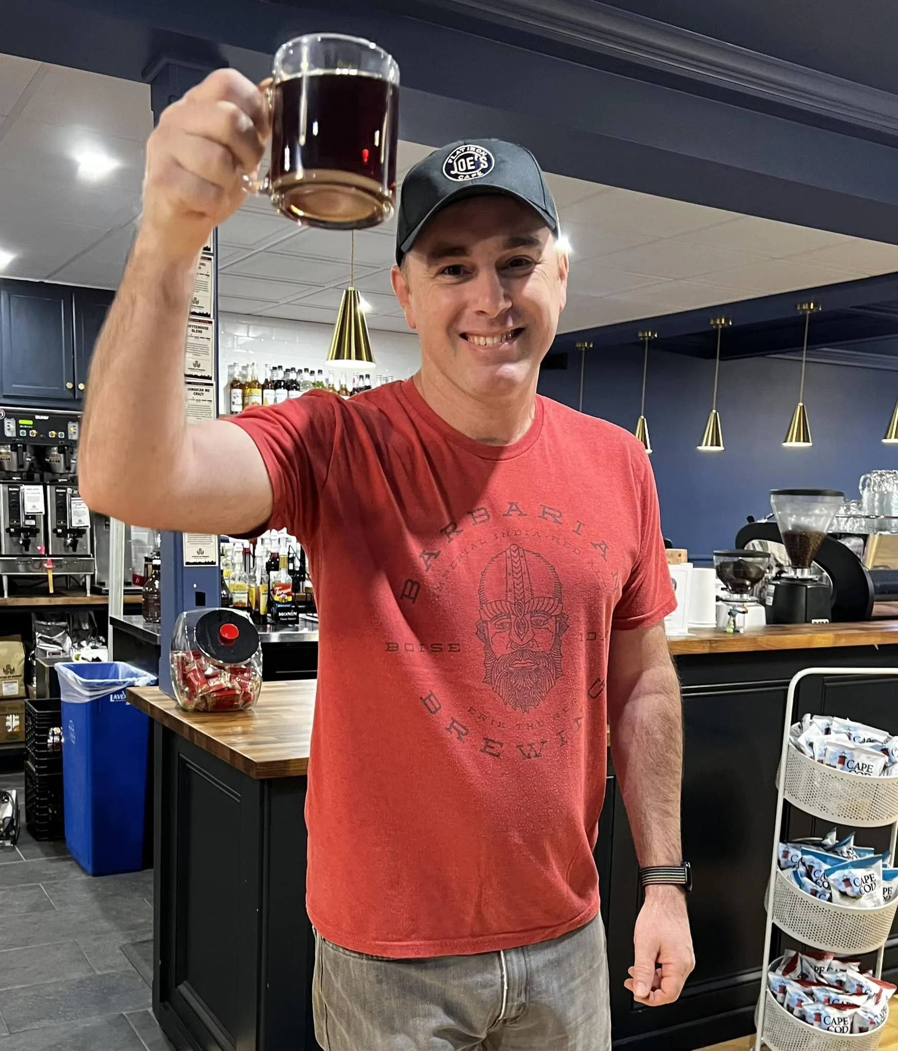 A man smiling and holding a glass mug filled with a dark beverage, standing in a coffee shop or cafe setting with shelves of bottles and coffee equipment in the background.