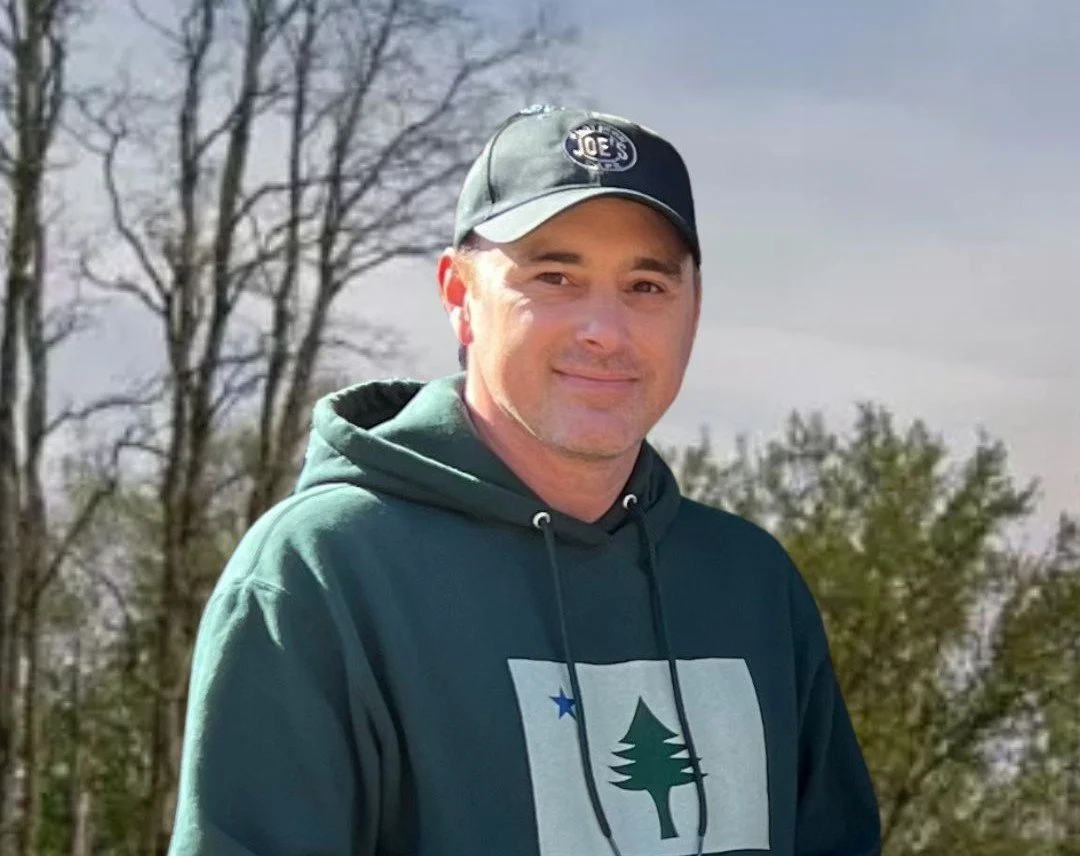 A man wearing a green hoodie and a gray baseball cap with the Joe's logo, standing outdoors during daytime with trees and a cloudy sky in the background.