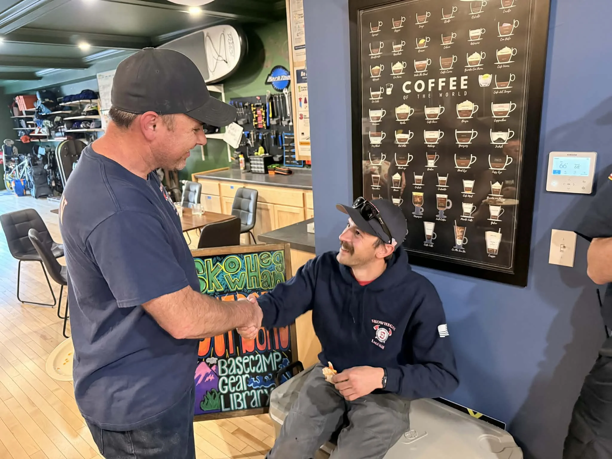 A man with a dark baseball cap interacting with a seated man holding food, in an indoor setting with a sign about coffee from around the world on the wall.