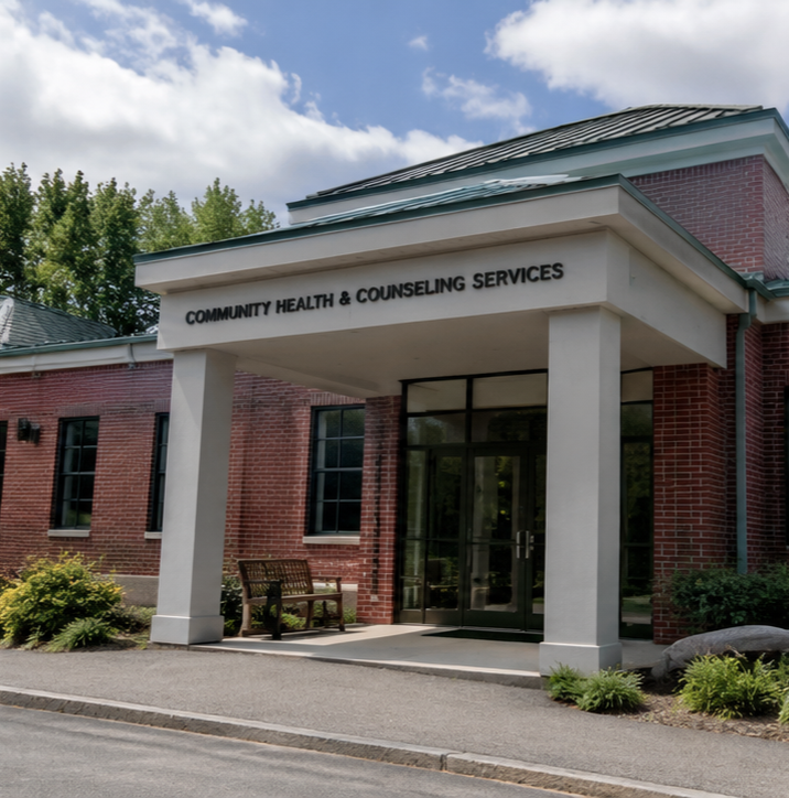 Exterior of a community health and counseling services building with a brick facade, glass doors, and a small porch with a bench, under a partly cloudy sky.