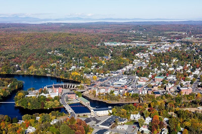 Aerial view of a small town with a river running through it, surrounded by trees with fall foliage, and mountains in the distance.