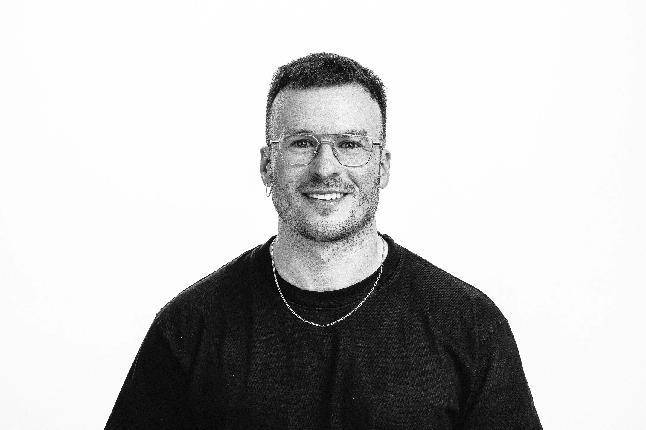 Black and white portrait of a smiling young man with glasses, earrings, wearing a black shirt and layered necklaces against a plain white background.