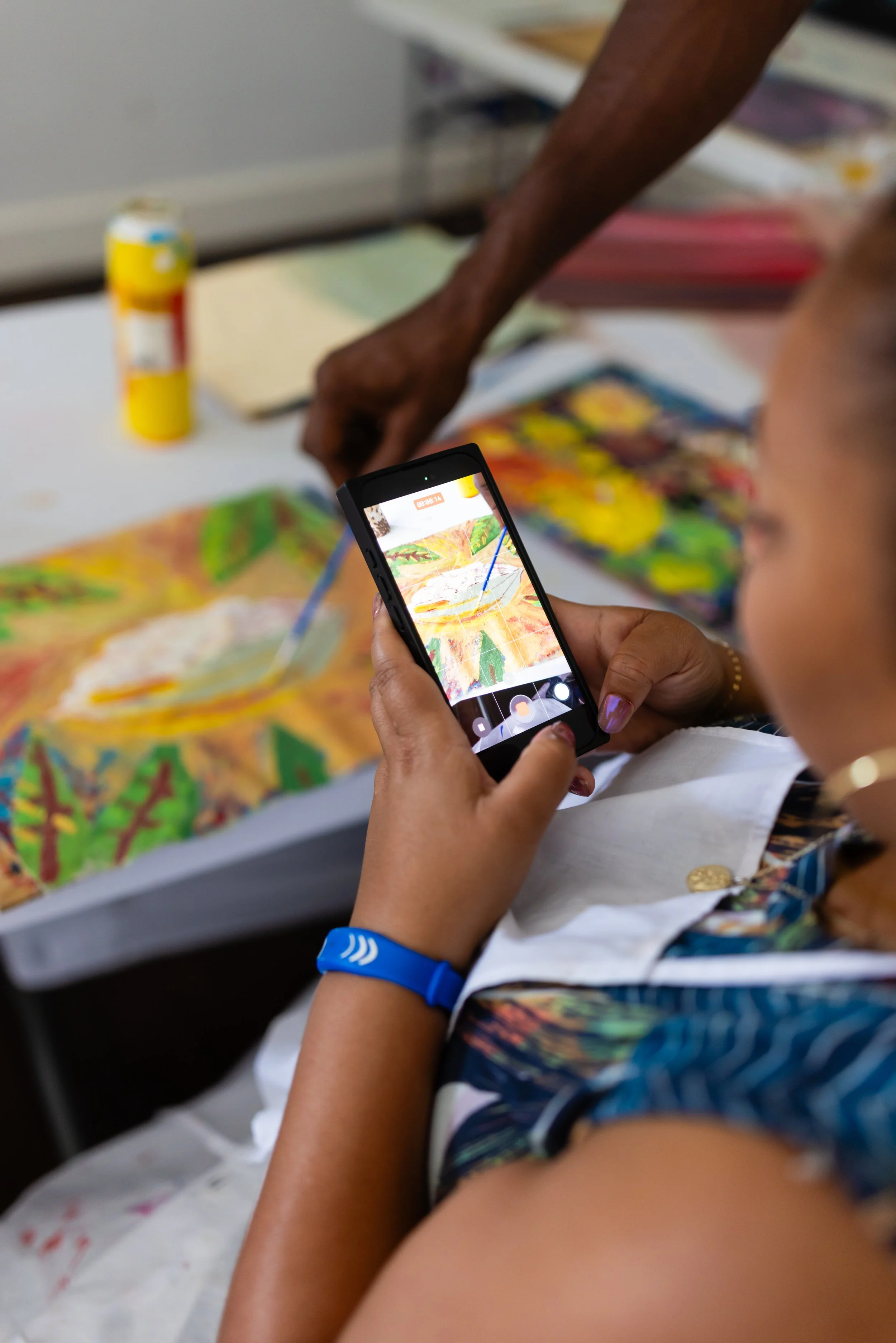 A person holding a smartphone taking a photo of colorful artwork on a table. The person's hand is visible, wearing a blue wristband.