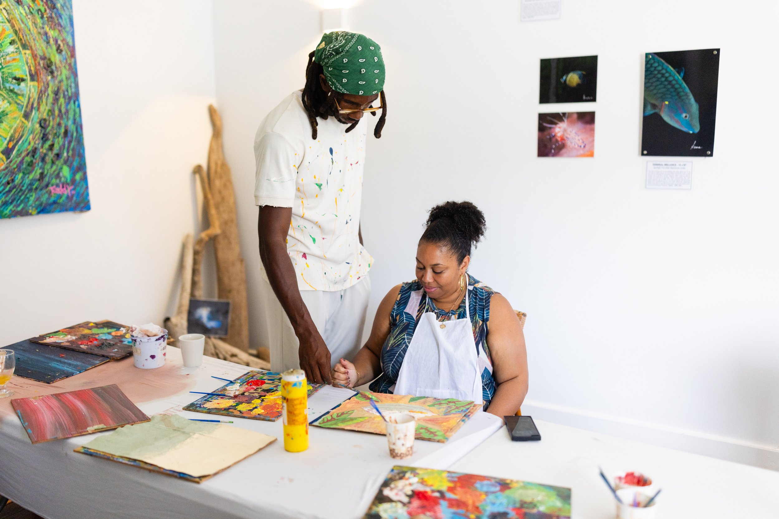 A man in a green bandana and glasses assisting a woman with curly hair in a blue and white striped dress, as she paints on a canvas at an art studio. The studio has artworks of fish and abstract paintings on the walls.