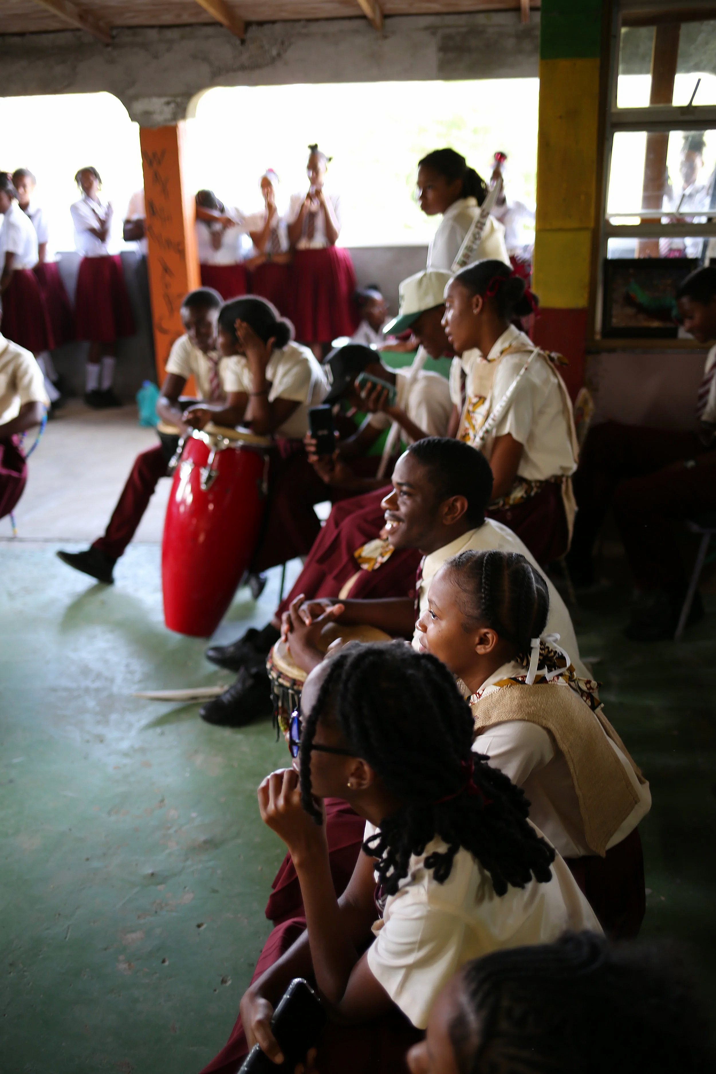 A group of school children sitting and standing in an indoor classroom with some holding musical instruments, including drums and flutes, with sunlight coming through open windows.
