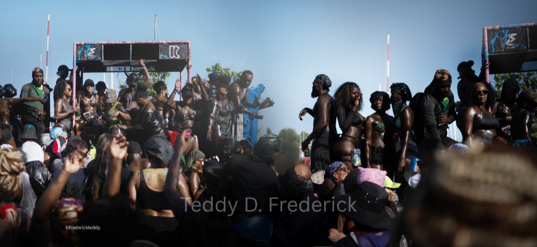 A large crowd of people, many covered in dark paint or mud, participating in an outdoor event or festival during daytime. Some are dancing on a stage while others are watching and enjoying the festivities.