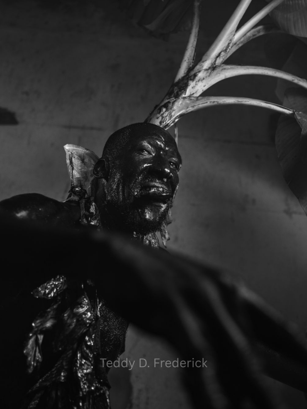 A black and white photo of an indigenous man with traditional body paint and adornments, standing next to a large plant with broad leaves.