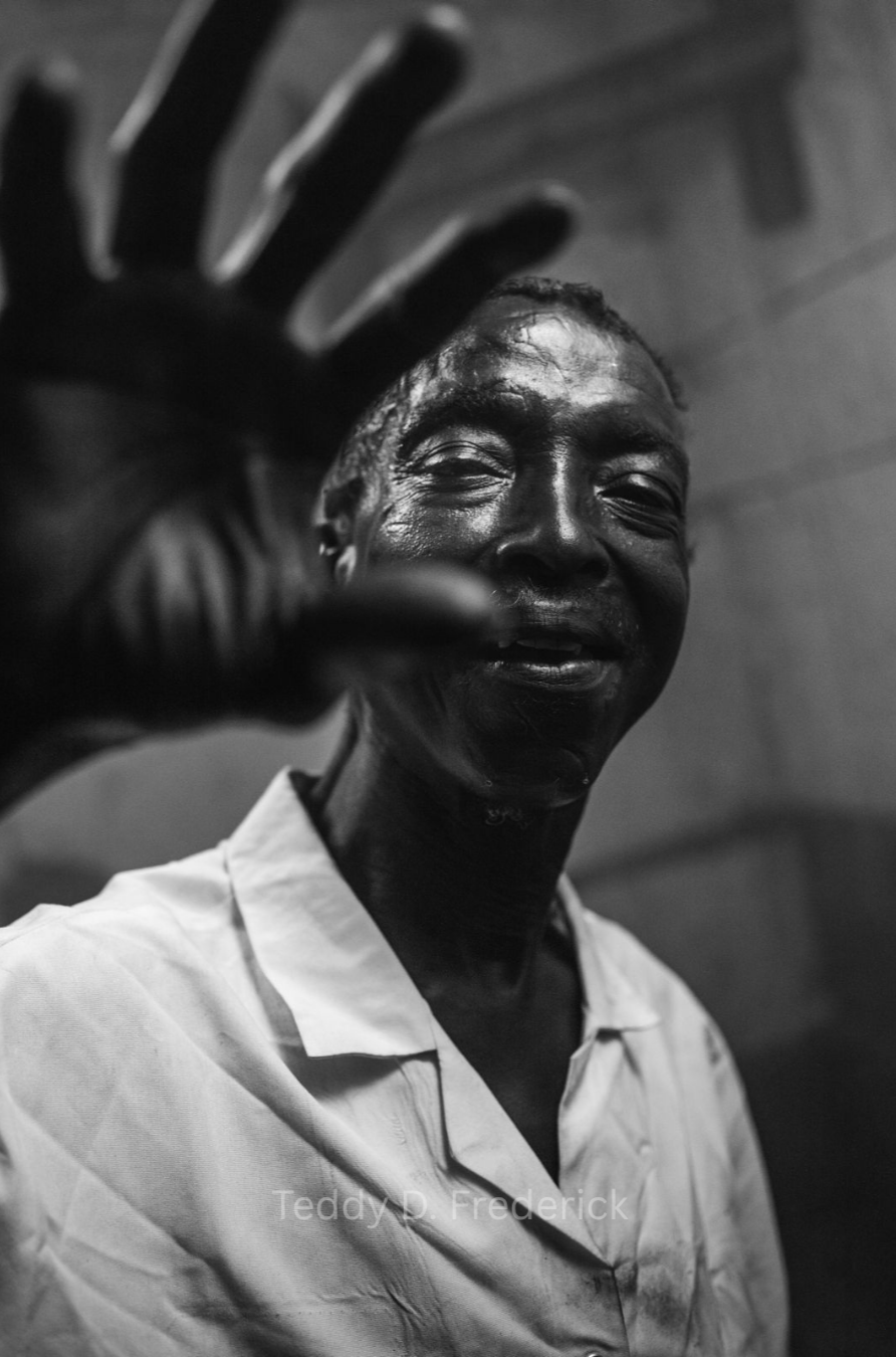 A smiling man with dark skin, raised hand reaching towards the camera, wearing a light-colored shirt, in black and white photography.