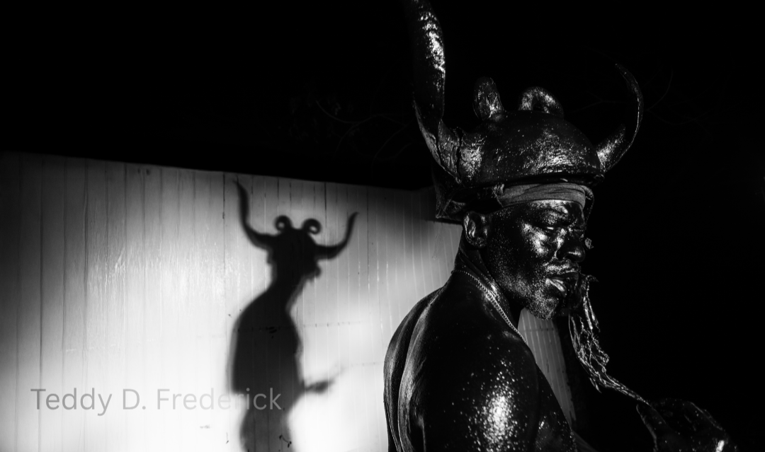 Black and white sculpture of a man wearing a large, elaborate headdress with curved horns, glasses, and a beard, with a prominent shadow of a horned figure cast on the wall behind him.