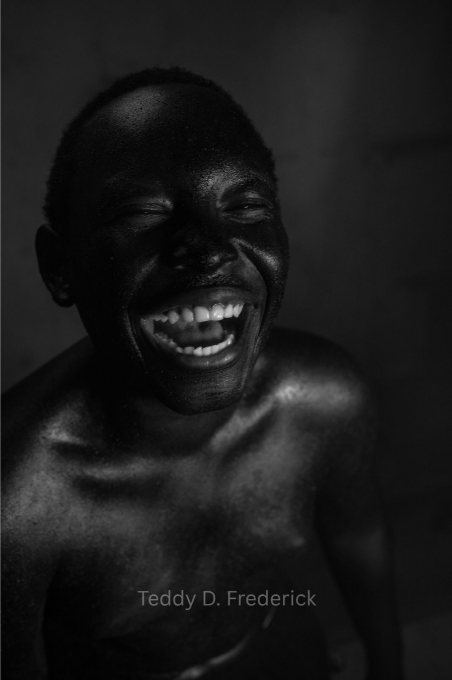 Black and white portrait of a man laughing with eyes closed, showing teeth and tongue, with a shiny, wet skin appearance.