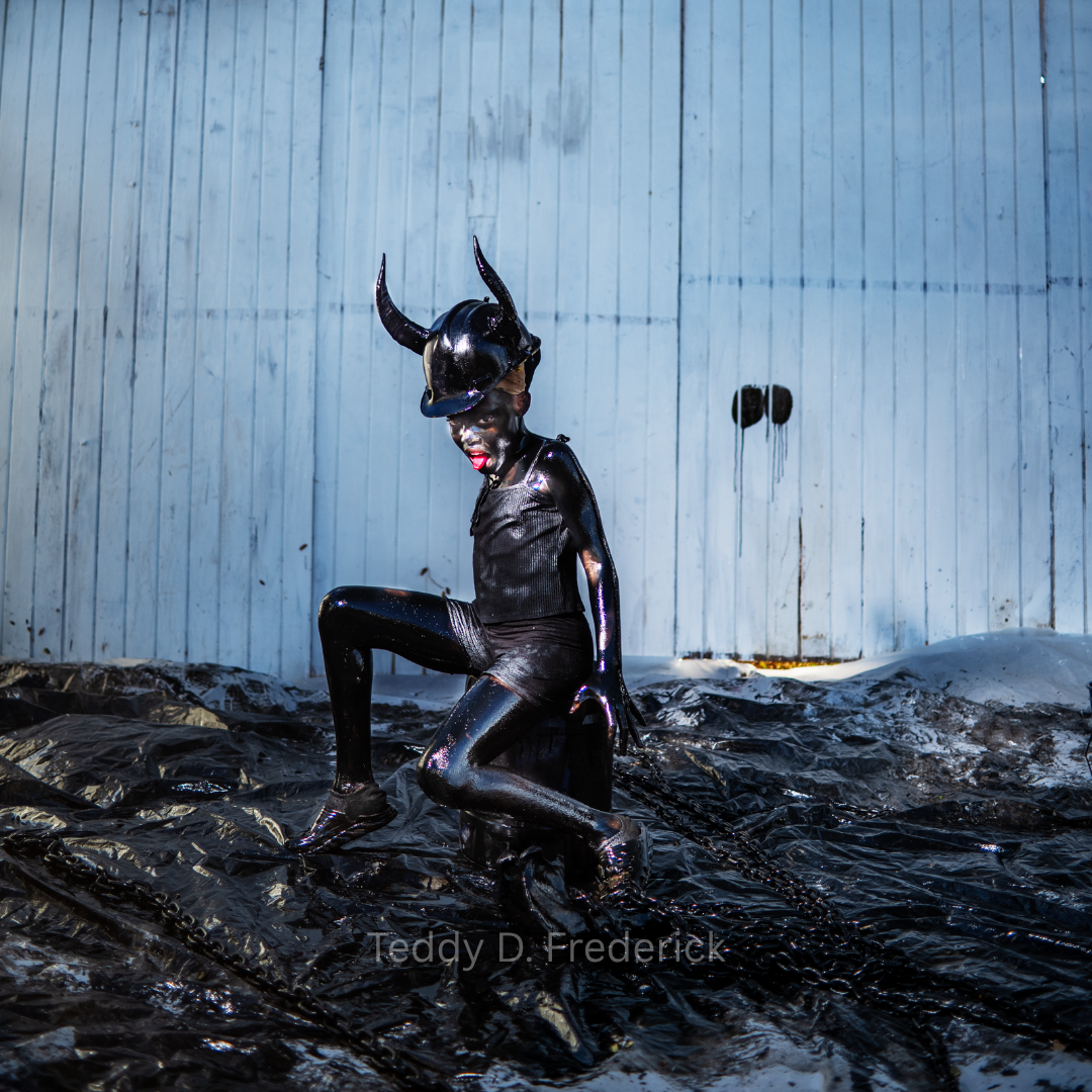 Person dressed in a shiny black outfit with a horned helmet, kneeling on a black plastic-covered surface in front of a light blue wooden wall.