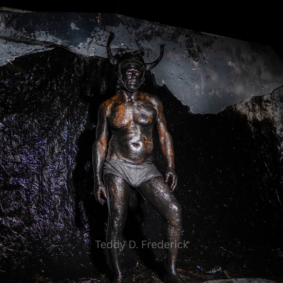 Man covered in black oil with an insect-like headpiece standing against a dark, textured backdrop.