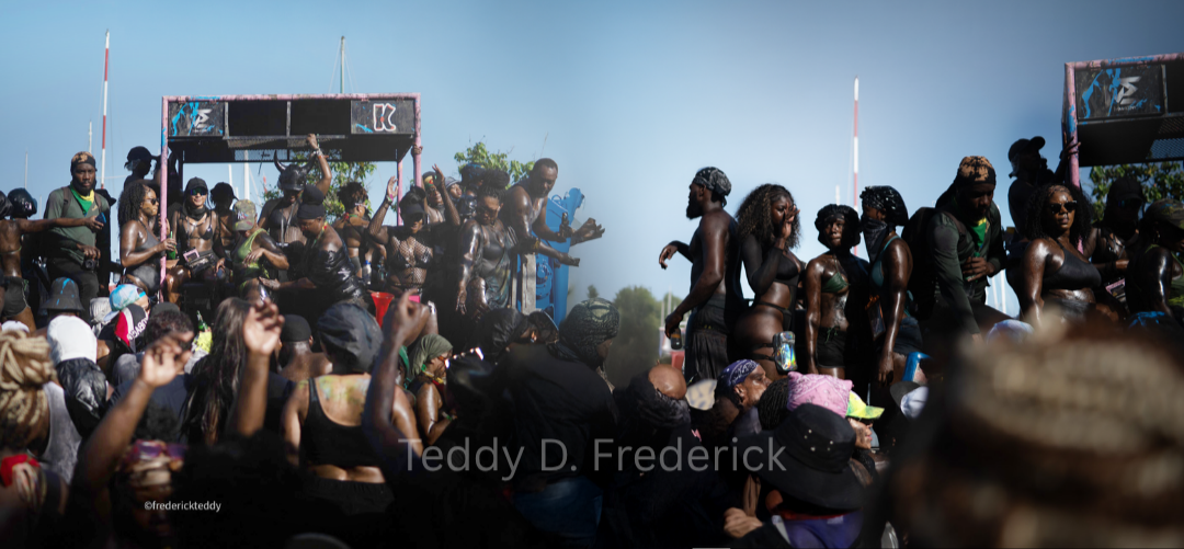 Crowd of people at an outdoor festival or music event, with some dancing and enjoying the sun. The background shows a stage structure and clear blue sky.
