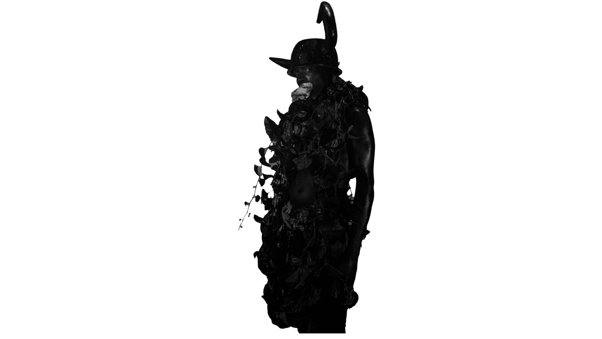 A person with black skin dressed in black, wearing a large black hat with a tall, curved peak, and adorned with black leaves and vines covering their chest and back, standing in front of a black background.