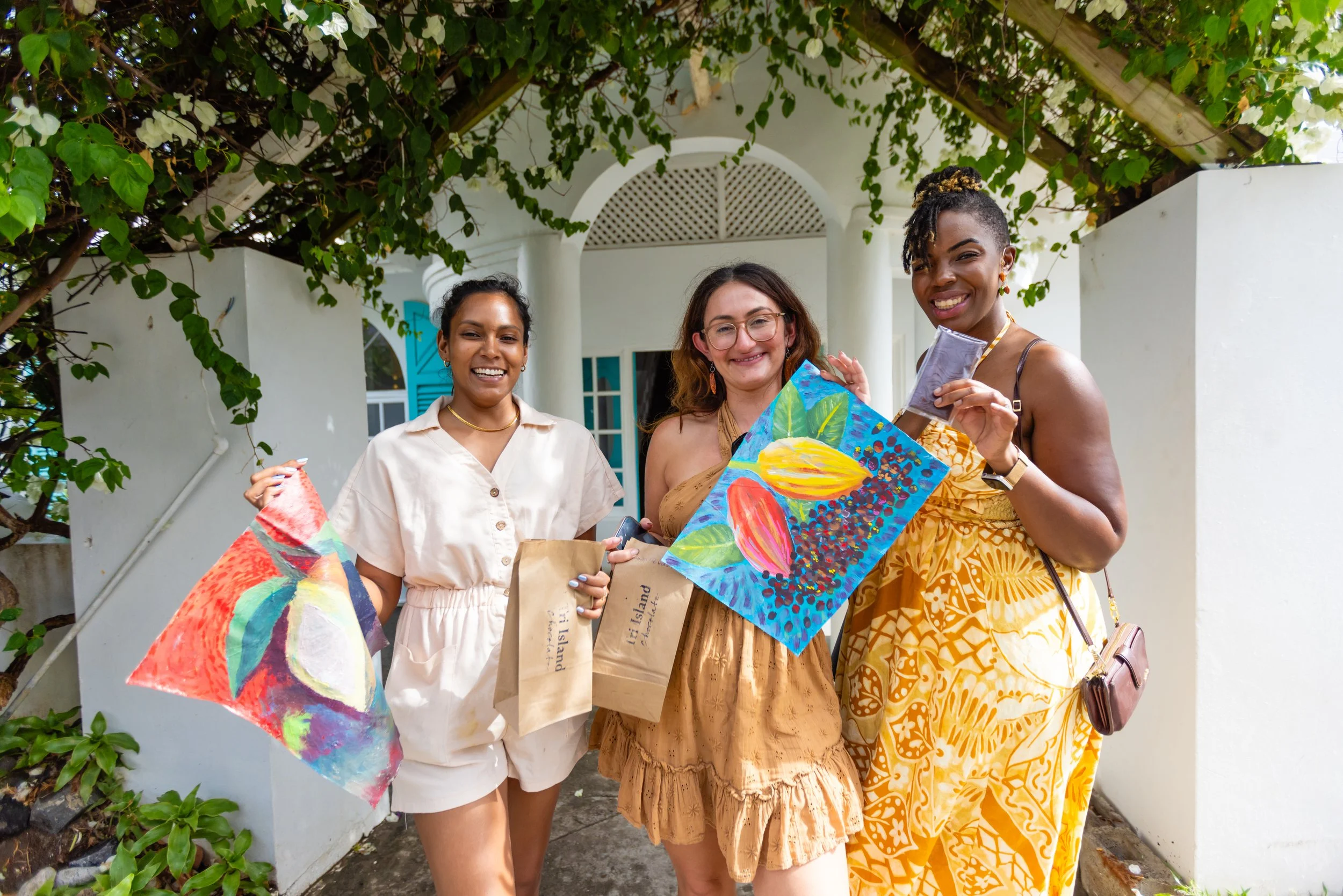 Three women smiling outdoors holding colorful paintings and gift bags, with greenery and a white house in the background.