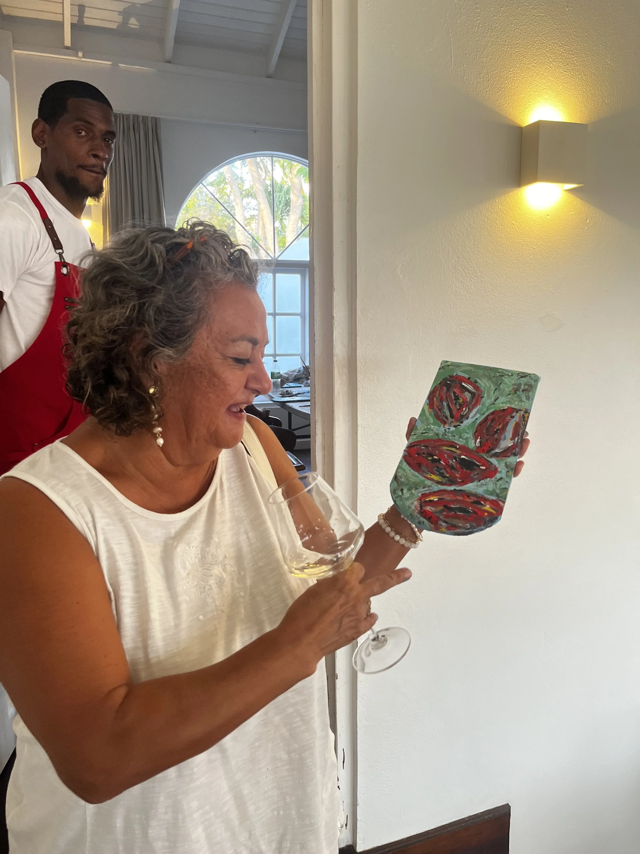 A woman with gray curly hair holding a wine glass and a colorful abstract painting, while a man in a red apron stands behind her in a bright, indoor setting with a large arched window and warm lighting.