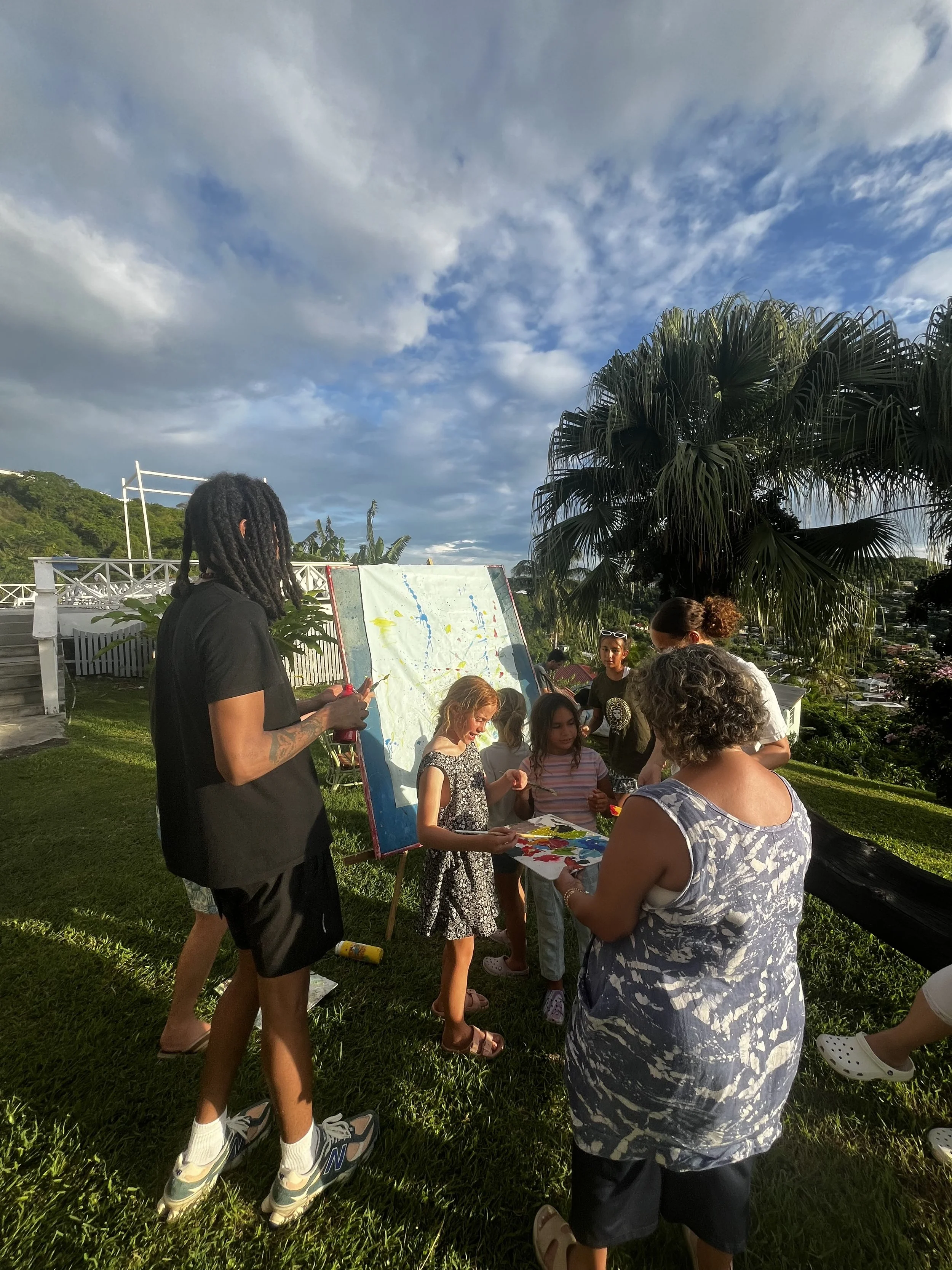 Group of people gathered outdoors, engaging in a painting activity on a sunny day with a partly cloudy sky, lush green grass, and tropical trees in the background.