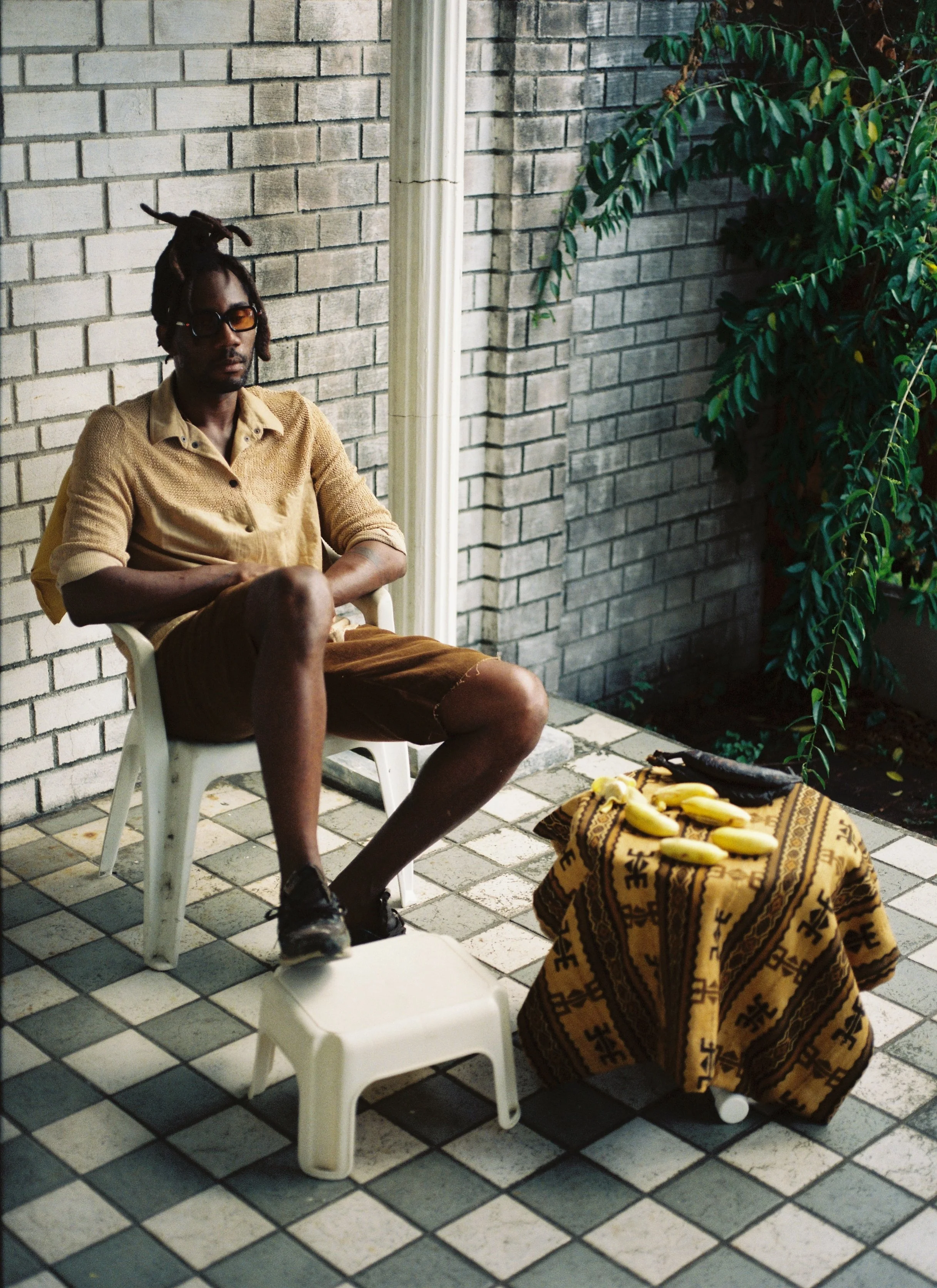 A man with dreadlocks and glasses sitting on a white plastic chair next to a small table with bananas on it, outside against a brick wall with a green leafy plant nearby.