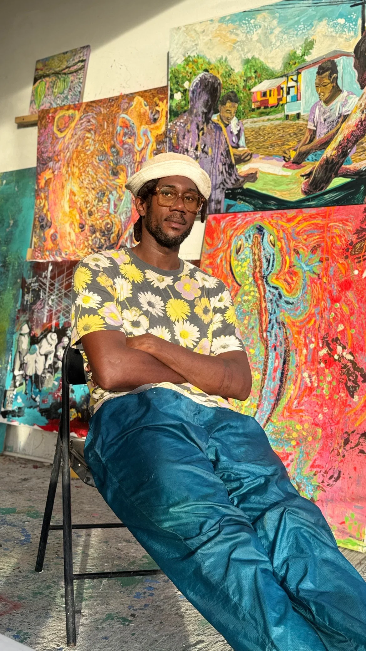 A man wearing a floral shirt, blue pants, glasses, and a white hat sitting on a chair in front of colorful abstract paintings in an art studio or gallery.