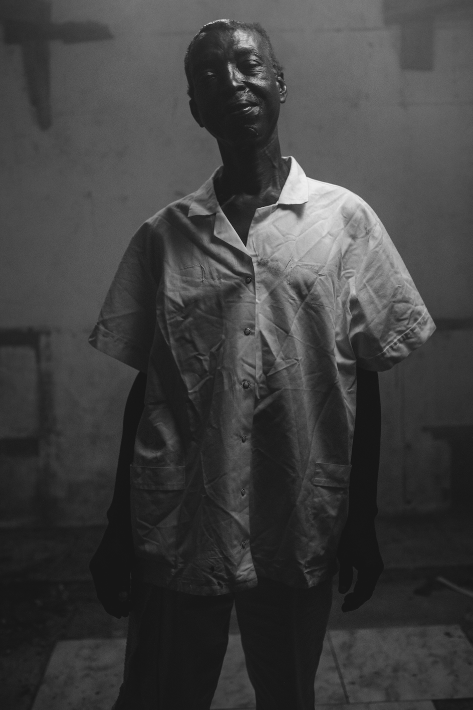 A black and white photo of an elderly person with short hair, wearing a wrinkled button-up shirt, standing indoors with a plain wall in the background.