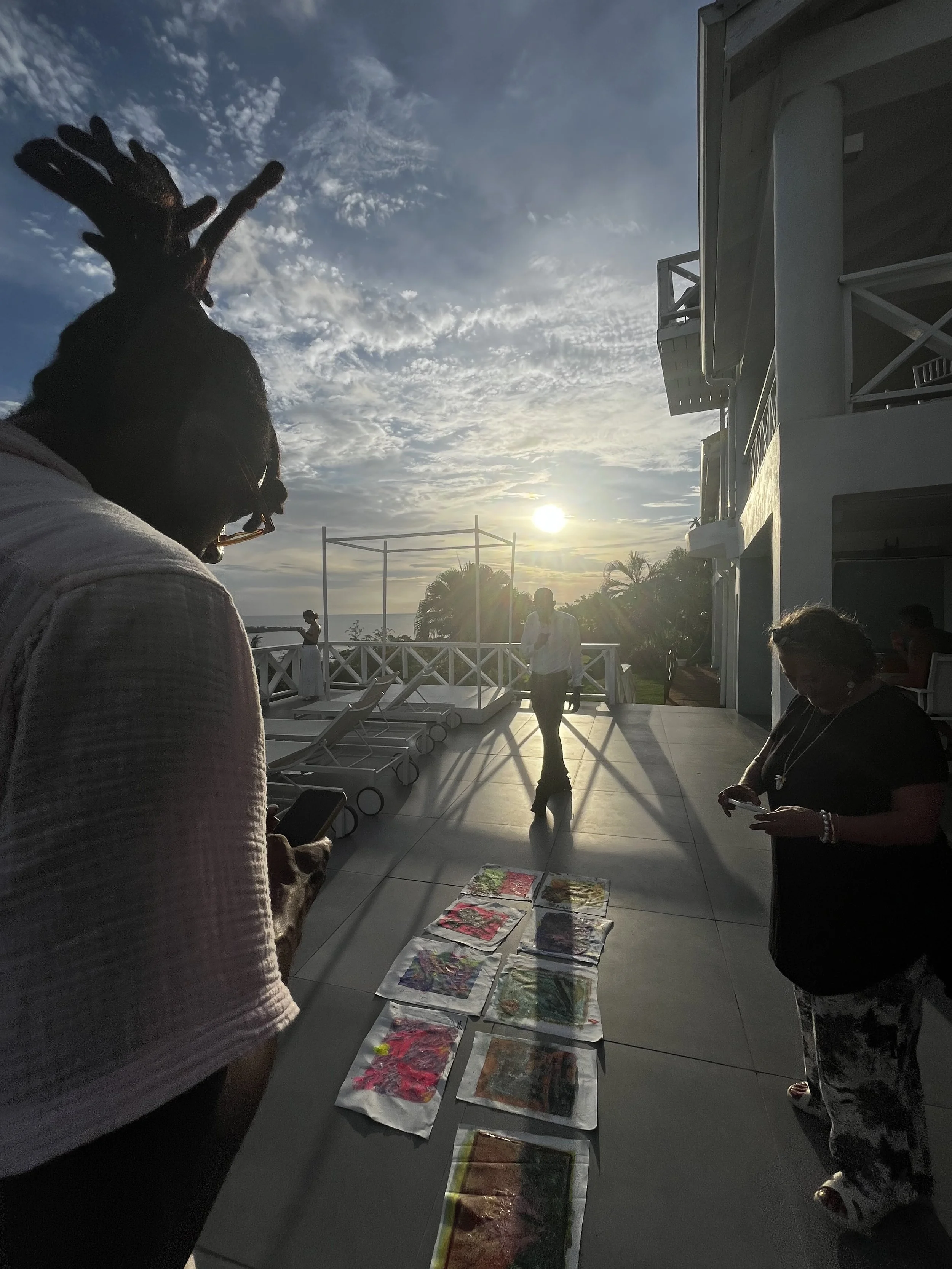 People standing on a tiled balcony or terrace, looking at artwork laid out on the ground. The sky is partly cloudy with the sun setting or rising in the background. Some individuals are using phones, and there are lounge chairs and a white railing visible.