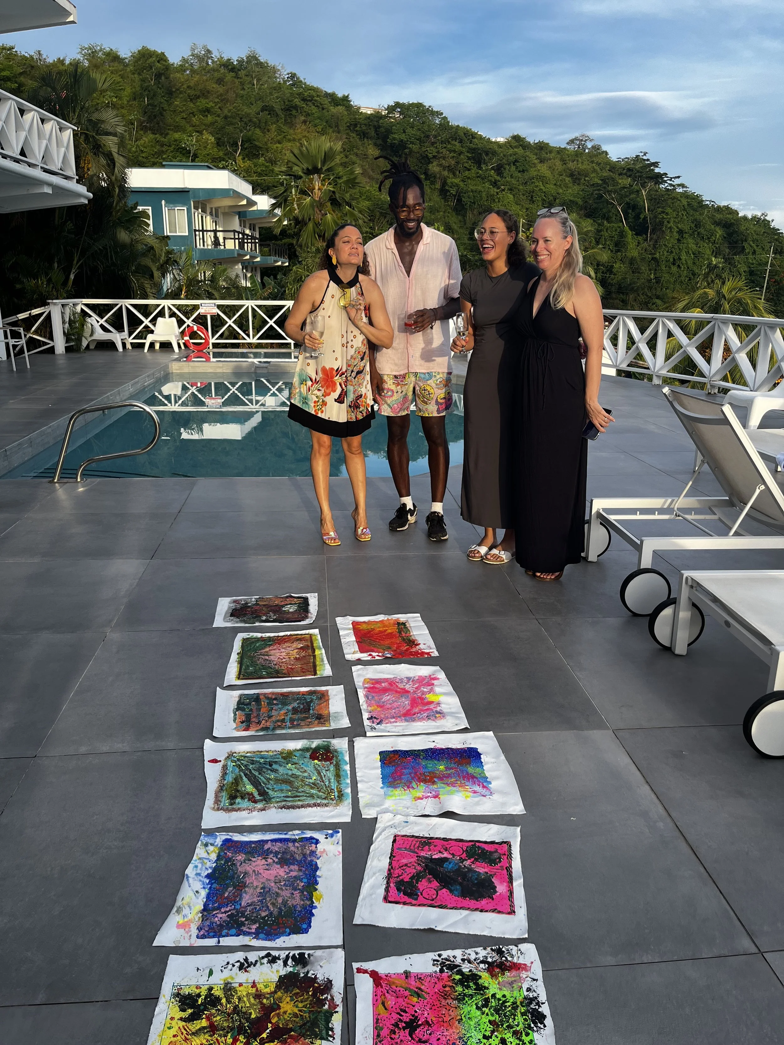 A group of four friends standing by a swimming pool, smiling and holding drinks at sunset. There are colorful abstract paintings laid out on the ground in front of them, with lush green hills and a partly cloudy sky in the background.