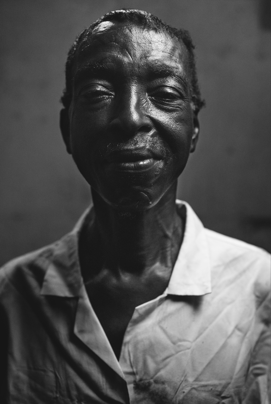 A close-up black and white portrait of an elderly man with short hair, smiling gently, wearing a light-colored collared shirt against a plain background.