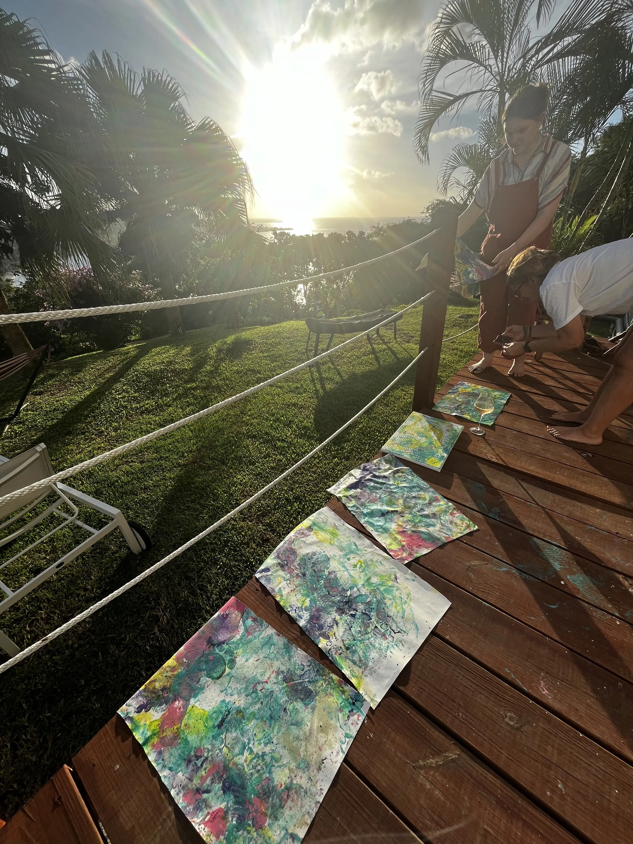 People engaging in an outdoor art activity on a wooden deck, with colorful paintings laid out, overlooking a lush green landscape and a bright setting sun.