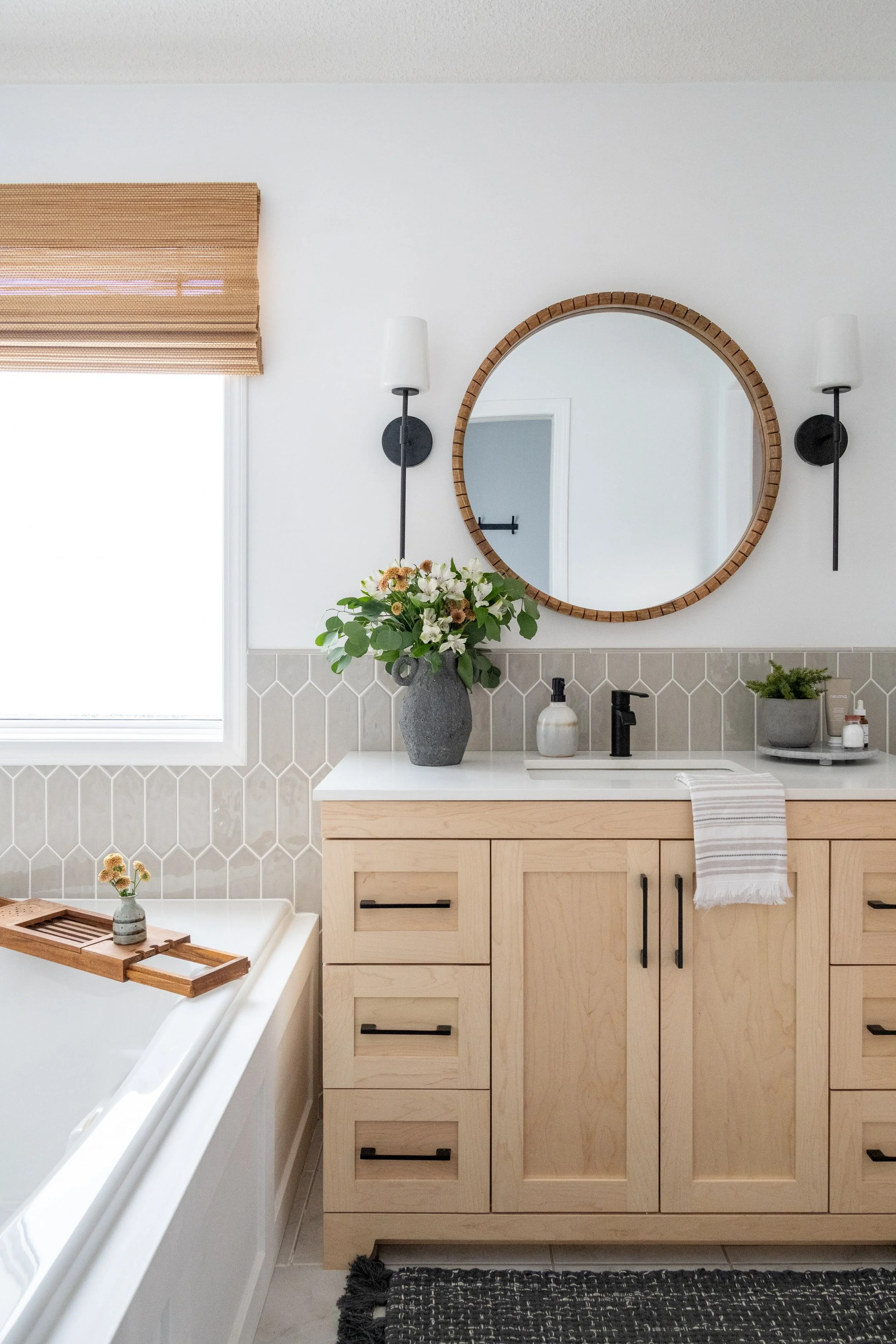 Modern bathroom with beige wooden vanity, round mirror, black wall sconces, and decorative plants.