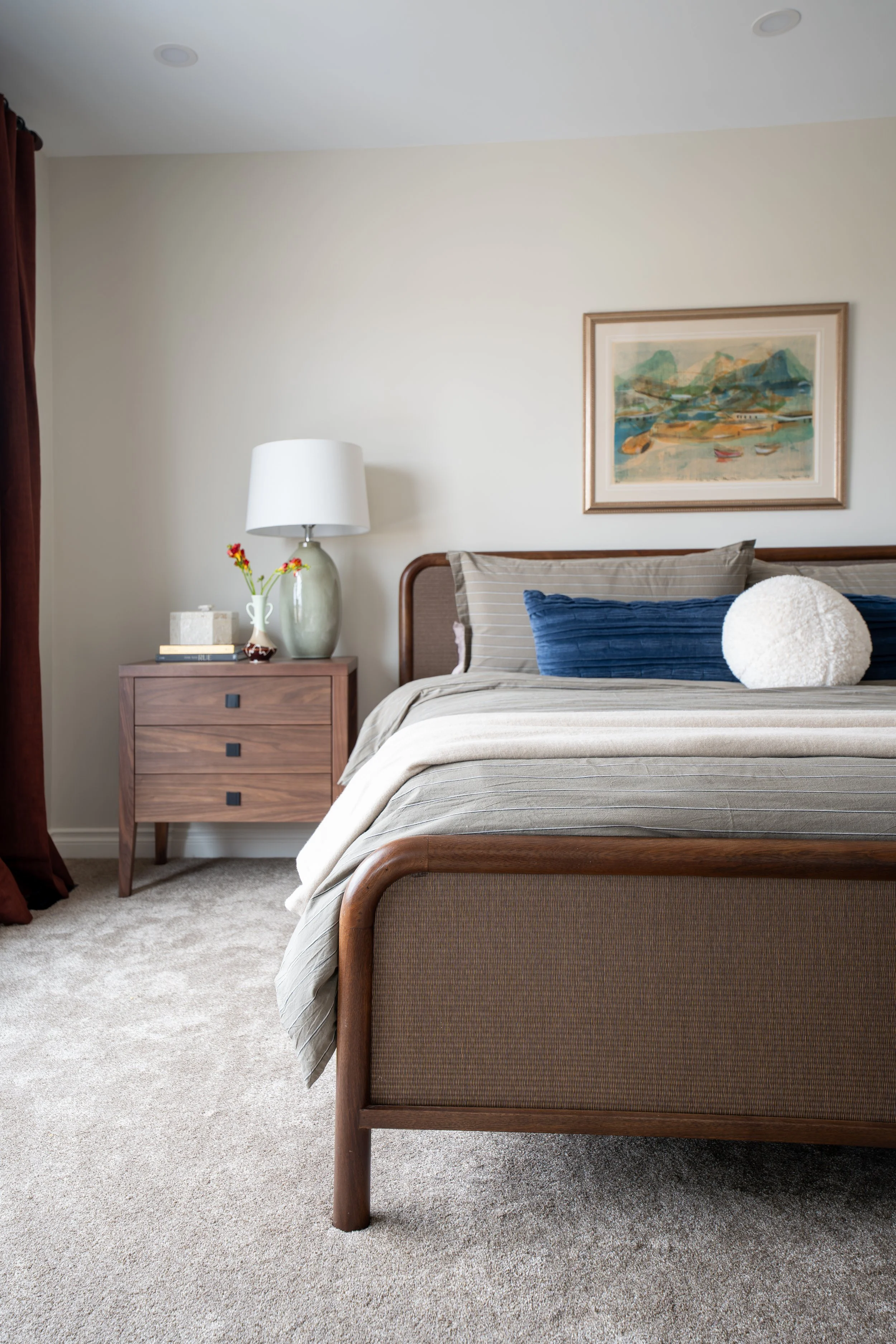 Spacious primary bedroom with warm toned wood and fabric bed frame with a walnut finish bedside table with a soft green table lamp and vintage vase and books and a painting above the bed