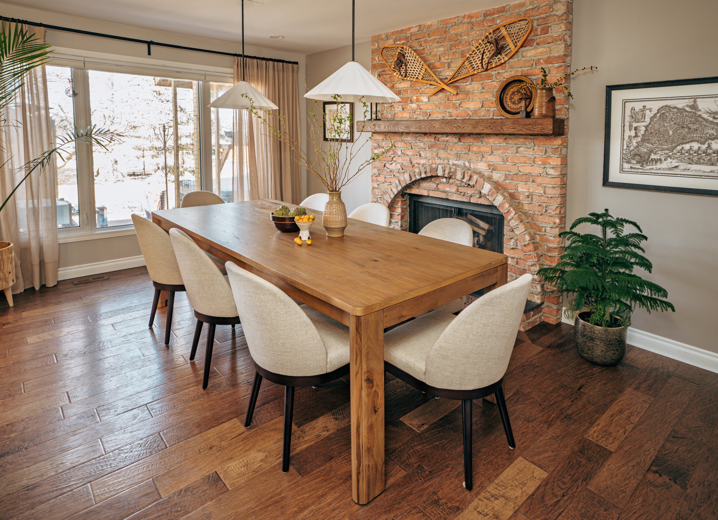 A dining room with a wooden table, eight cream-colored upholstered chairs, a brick fireplace with vintage snowshoes and a woven basket on the mantel, potted plant near the fireplace, framed artwork on the wall, large windows with beige curtains, and hardwood floors.