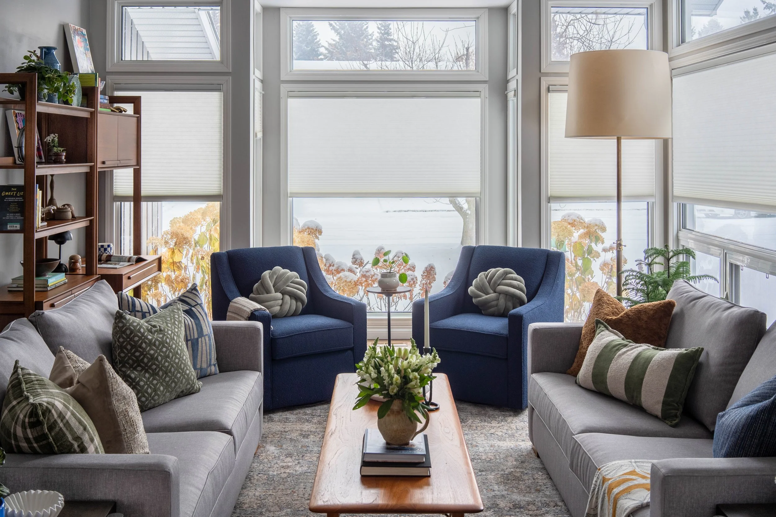 A cozy living room with two gray sofas and two blue armchairs. The sofas have various patterned pillows, and the armchairs have knotted pillows. There is a wooden coffee table with a flower arrangement and books. Large windows with white blinds let in natural light, revealing a snowy landscape outside, with trees and a house roof. A tall floor lamp and a wooden bookshelf filled with books and decorative items are also visible.