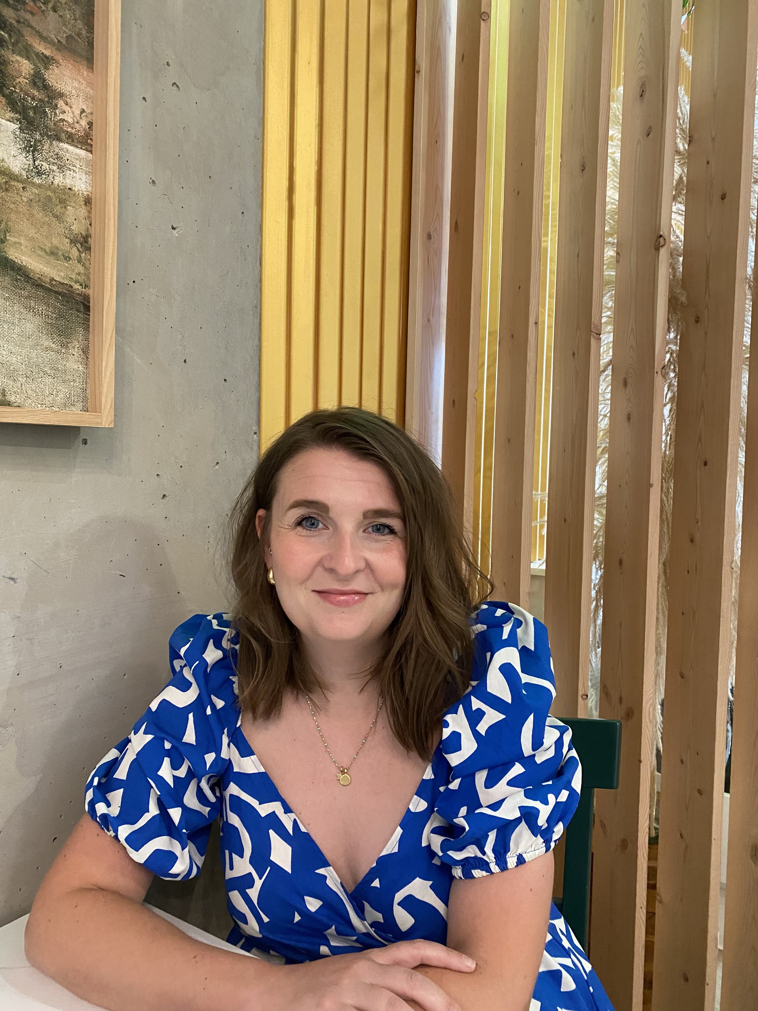 A woman with shoulder-length brown hair smiling at the camera. She is wearing a blue and white patterned dress and gold jewelry. She is sitting at a table in a modern indoor setting with wooden panel decor and a concrete wall background.