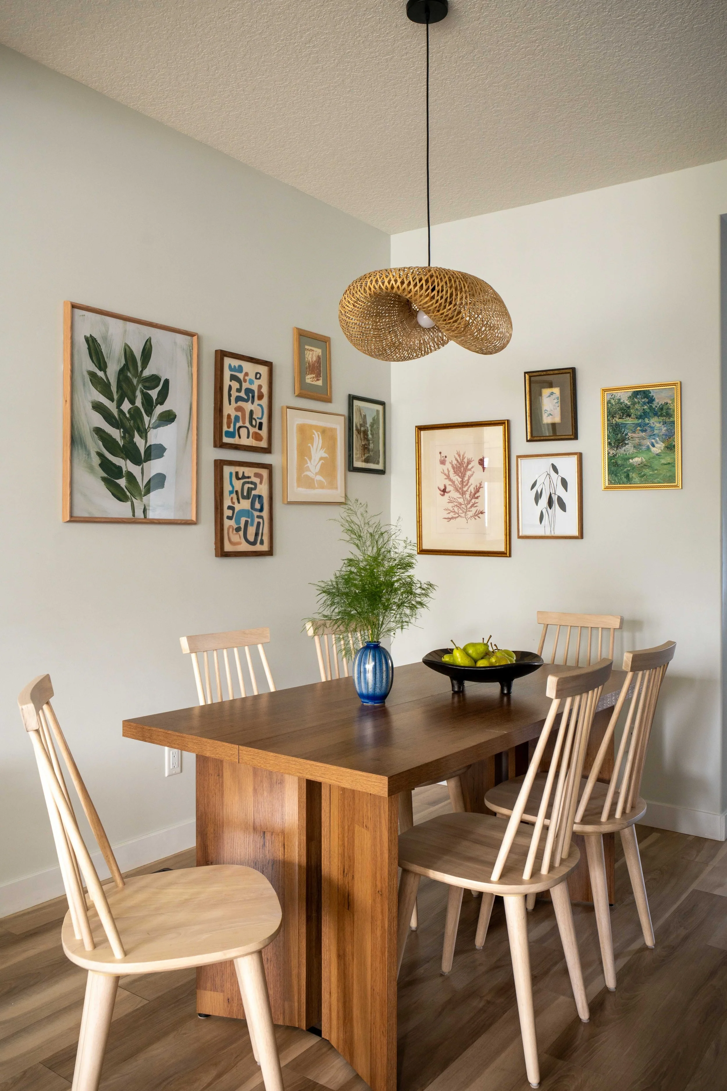 Dining room with wooden table, six light wood chairs, a blue vase with green foliage, a black bowl filled with green apples, and framed artwork on the wall. Hanging light fixture with a woven, organic design.
