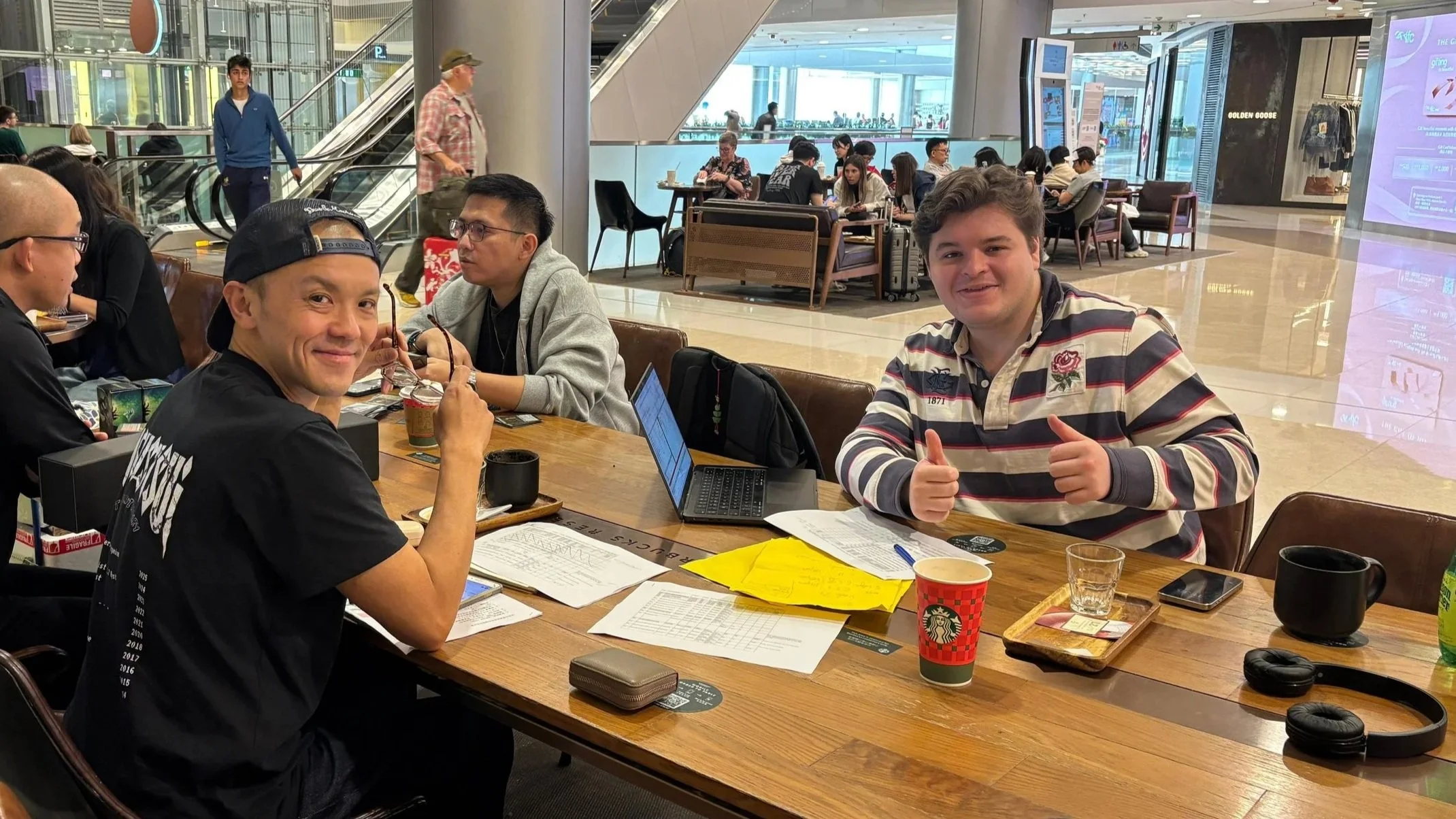Two young men sitting at a table in a busy airport lounge, smiling and giving thumbs up. The table has papers, a laptop, drinks, and headphones. Other travelers are visible in the background.