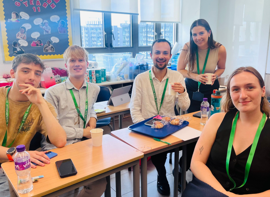 Five young adults sitting and standing around a table in a classroom, smiling. On the table are snacks, drinks, a tablet, and a notepad. Behind them, windows let in natural light, revealing a cityscape of tall buildings. A bulletin board on the wall 