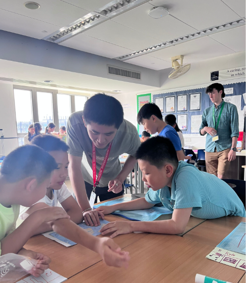 Students and teacher working together on a classroom project at a table with books and papers, others observing in the background.