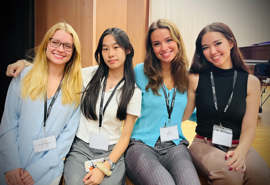 Four young women sitting together, smiling, at a conference or event, wearing name badges and lanyards.