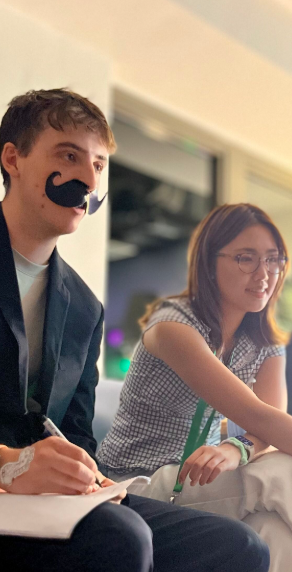 Two young adults sitting at a table, one wearing a fake mustache and holding a pen, the other wearing glasses and smiling.