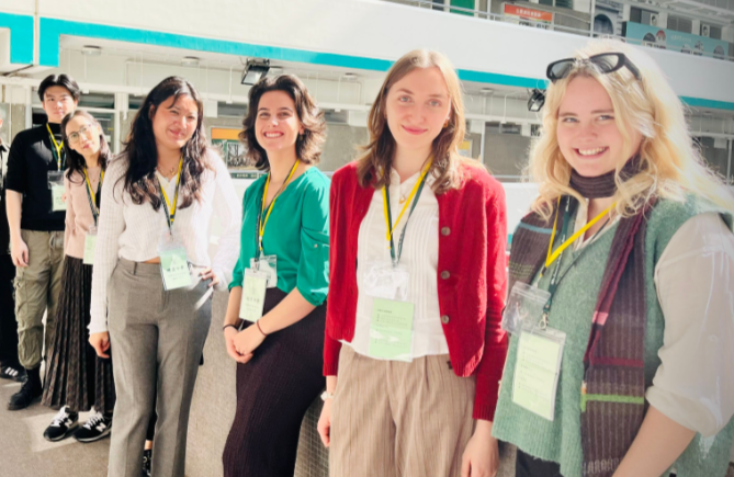 Group of five women and two men standing in a line at an indoor conference, wearing name badges and casual business attire, smiling at the camera.