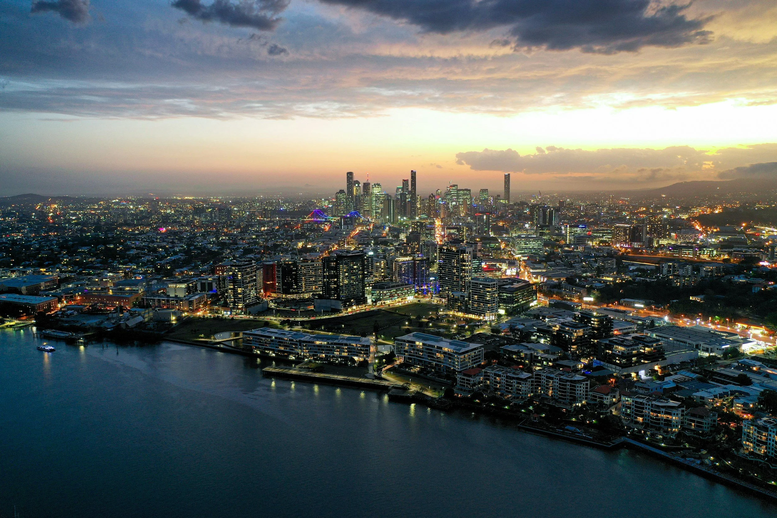 Aerial view of a city skyline at dusk with illuminated buildings, water in the foreground, and a cloudy sky.