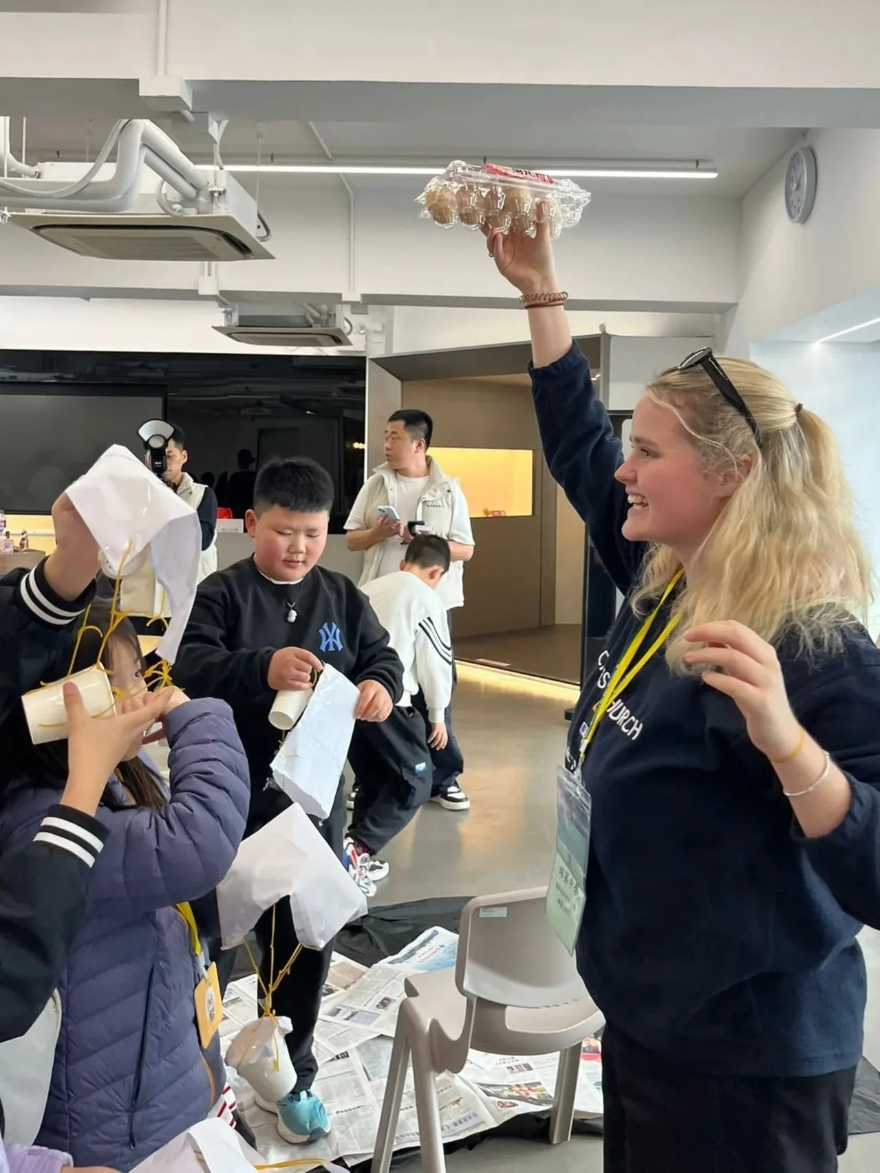 A young woman with blonde hair, wearing sunglasses and a Navy-blue jacket, is holding a carton of eggs overhead. Several children are gathered around, some with eggs and paper, inside a modern, well-lit indoor space.