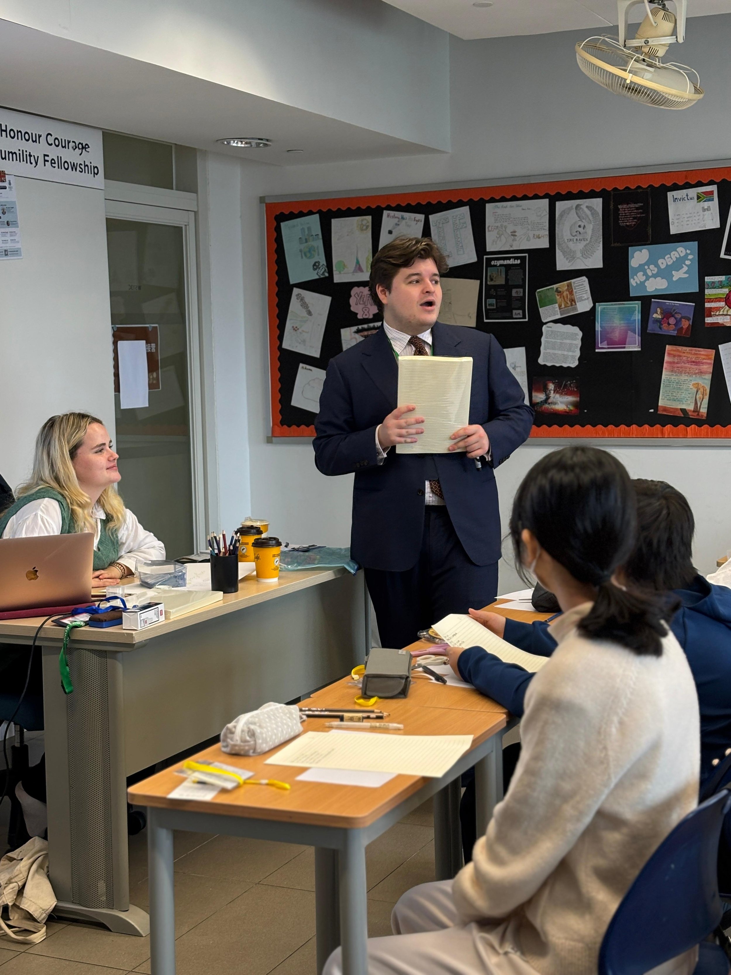 A classroom scene with students and a teacher. The teacher, standing and holding papers, appears to be giving a presentation. Seated students are taking notes and listening. Decorated bulletin board in the background displays various posters and artw