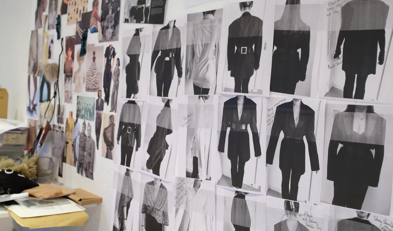 Wall with black and white photos of various fashion designs and clothing sketches, along with notes and small images, likely in a fashion design studio.